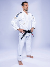 A man in a SEKAI 3.0 BJJ Gi White Navy and a black belt stands barefoot with hands on hips, gazing to the side against a light background.