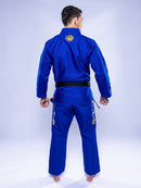 A person wearing the SEKAI 3.0 BJJ Gi Blue Yellow with a black belt stands barefoot, facing away to display the back of the uniform, which features patches and logos on both the jacket and pants.