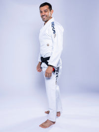A man wearing the SEKAI 3.0 BJJ Gi White Navy with dark lettering on the sleeve and pants stands barefoot on a white background, smiling and looking at the camera.