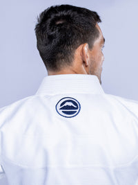 A man with short dark hair is seen from behind wearing the SEKAI 3.0 BJJ Gi White Navy, which features a circular black and white logo with two wave shapes on the back of the collar.