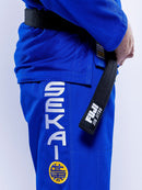 A person wearing the SEKAI 3.0 BJJ Gi Blue Yellow, featuring “SEKAI” and a yellow patch on the pants, holds a black Fuji Jiu Jitsu belt. The photo captures their torso and arm against a light background.
