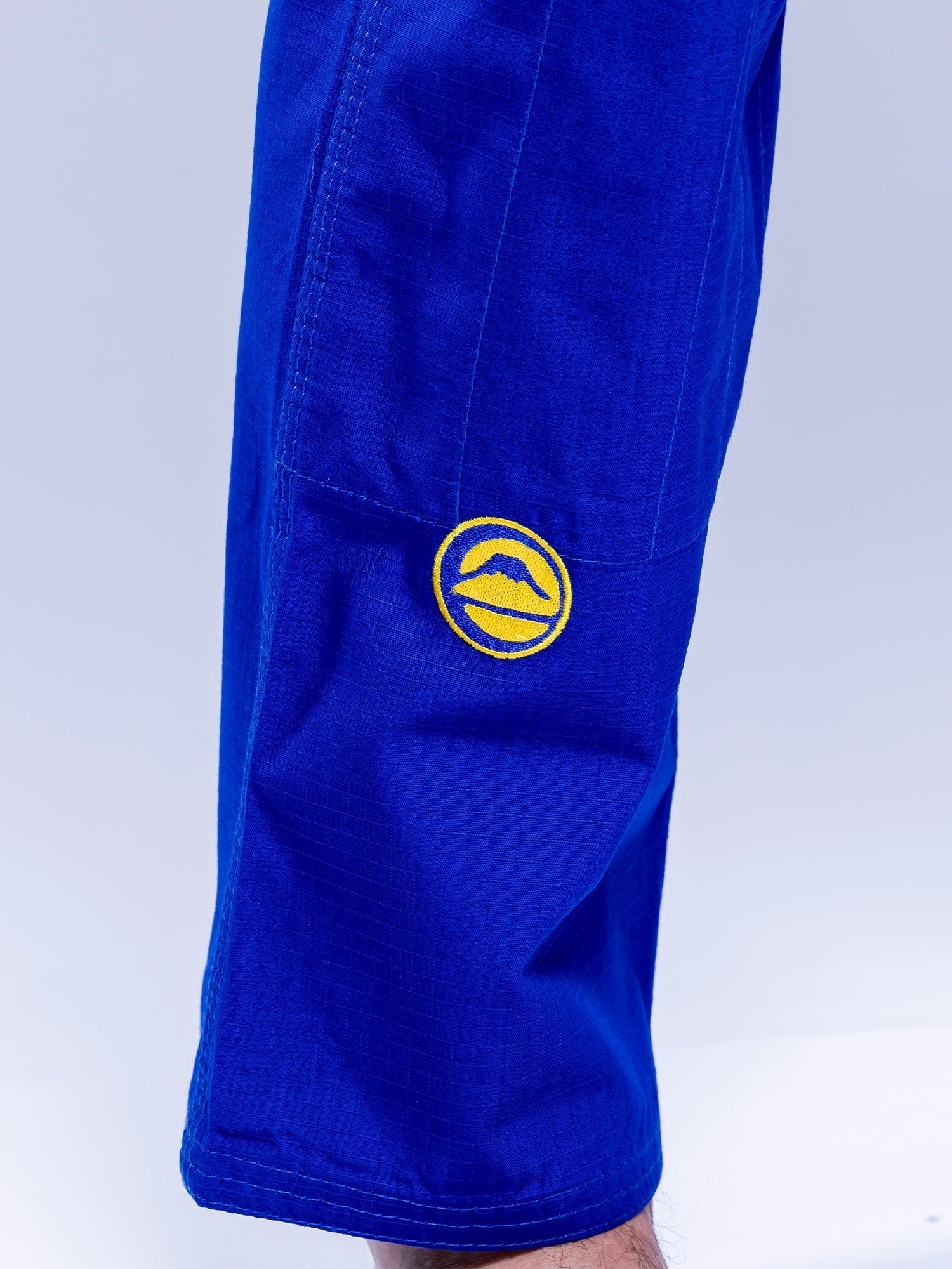 A close-up of someone wearing the SEKAI 3.0 BJJ Gi Blue Yellow, featuring a yellow circular mountain patch on the upper leg. The background is light and uncluttered.