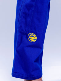 A close-up of someone wearing the SEKAI 3.0 BJJ Gi Blue Yellow, featuring a yellow circular mountain patch on the upper leg. The background is light and uncluttered.