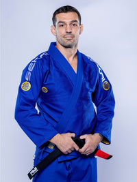 A man wearing the SEKAI 3.0 BJJ Gi Blue Yellow with patches stands facing forward, holding a black belt with a red stripe, against a plain light background.