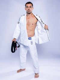 A man stands barefoot on a white floor wearing the SEKAI 3.0 BJJ Gi White Navy with patches, holding a black belt in his left hand and opening his jacket with his right to reveal his chest.
