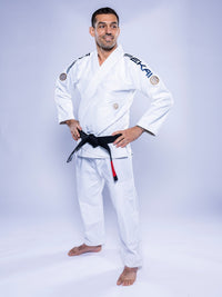 A man stands barefoot on a plain light background, smiling with his hands on his hips and wearing the SEKAI 3.0 BJJ Gi White Navy with a black belt.