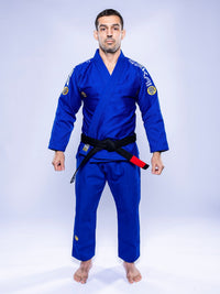 A man stands barefoot on a white background, wearing the SEKAI 3.0 BJJ Gi Blue Yellow with a black belt, facing forward and arms relaxed at his sides.
